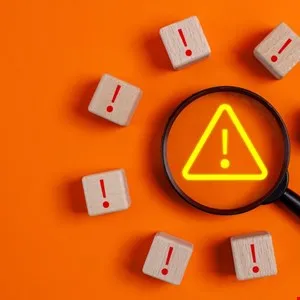 Magnifying glass over warning sign surrounded by exclamation point blocks.