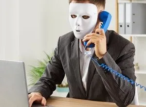 Businessman wearing a white mask talks on a vintage blue phone while working on a laptop.