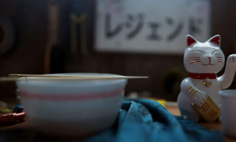 A white Maneki-neko cat figurine sits next to a bowl of noodles with chopsticks.