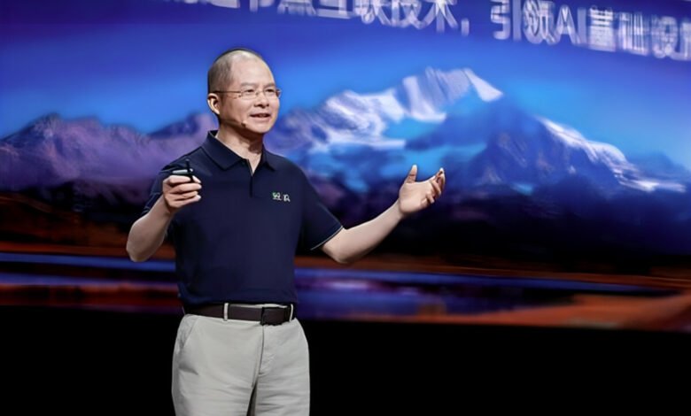 A man in a dark polo shirt gestures during a presentation with a mountain backdrop.