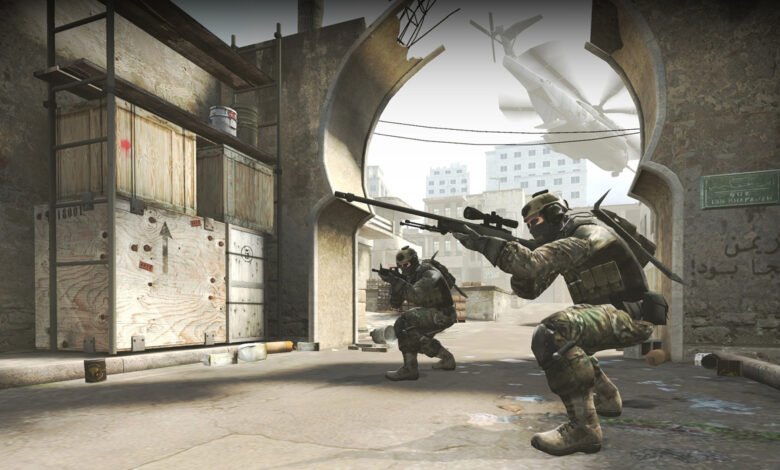 Two soldiers in camouflage gear take aim in a war-torn city street, a helicopter visible in the background.