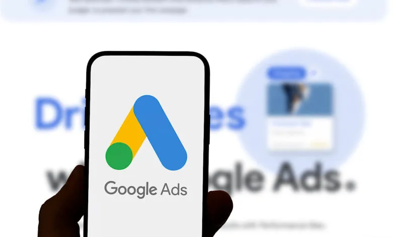 Close-up of a smartphone displaying the Google Ads logo. The background shows a blurred Google Ads webpage.
