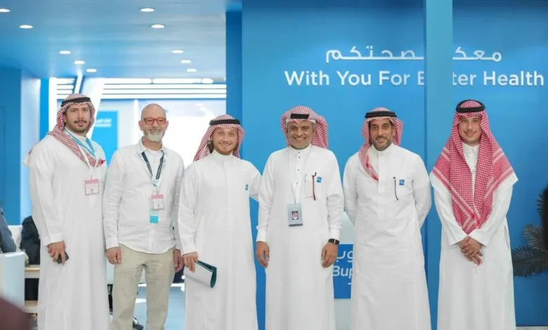 Group of men in traditional Middle Eastern attire and business casual pose in a modern office with blue branding.