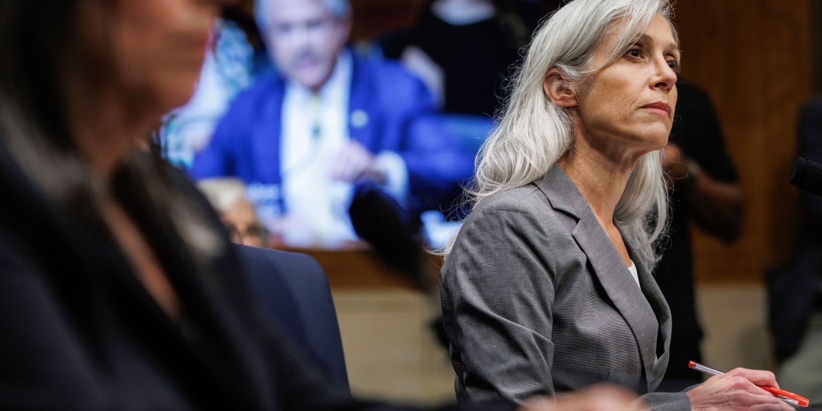 A woman with gray hair in a gray suit sits at a table, taking notes during a hearing or meeting.