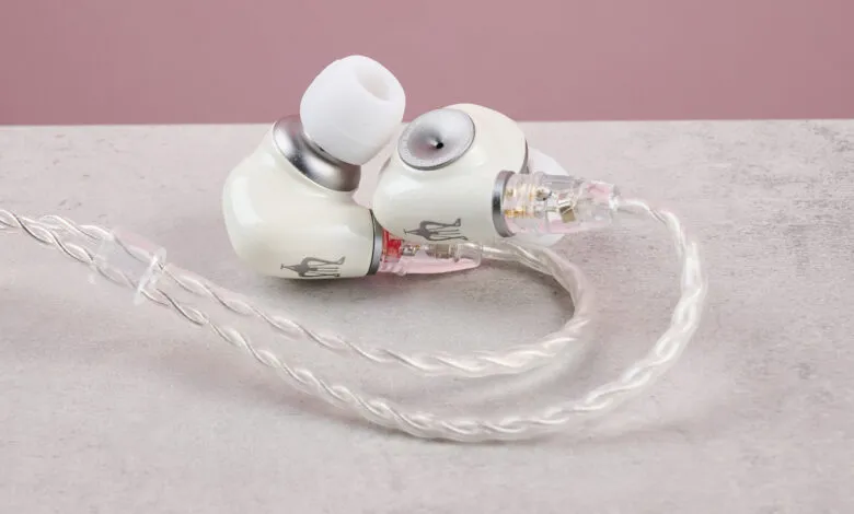 Close-up of Meze Audio Alba in-ear monitors, off-white with silver accents, resting on a light gray surface.