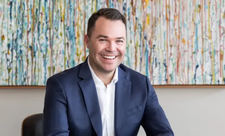 Aaron Thorne smiles broadly in a navy suit and white shirt, seated in front of abstract art.