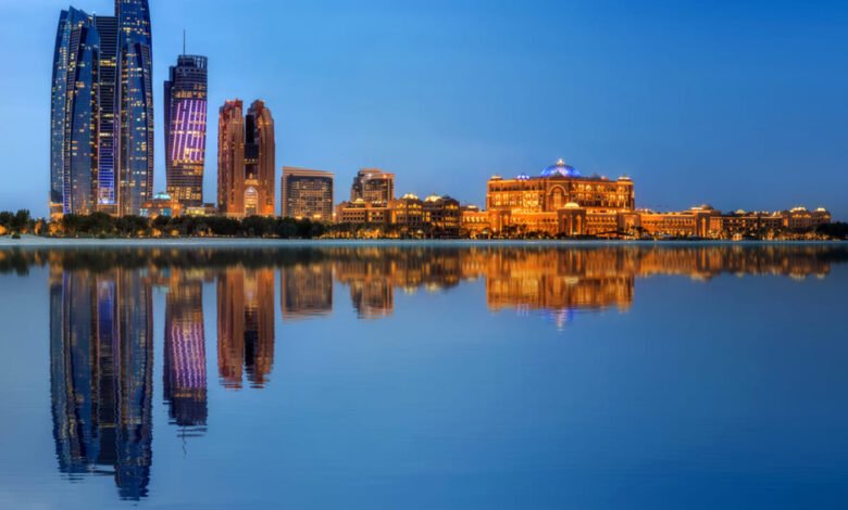Skyline of Abu Dhabi with modern skyscrapers and the illuminated Qasr Al Watan palace reflected in water.