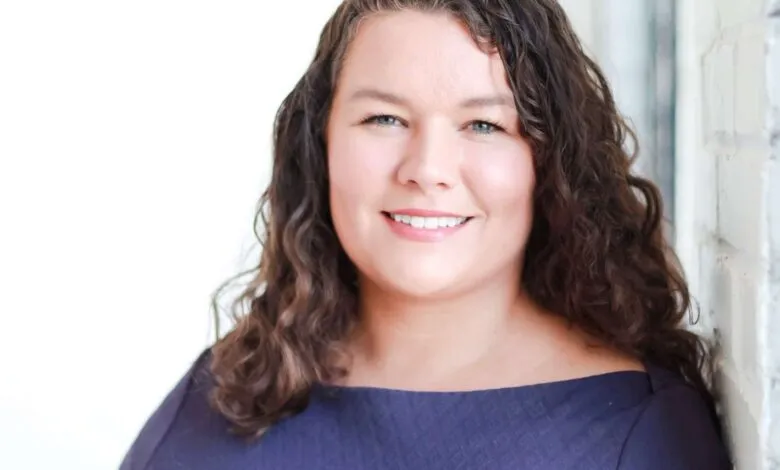 Smiling woman with curly brown hair, wearing a dark purple dress, leans against a white brick wall.