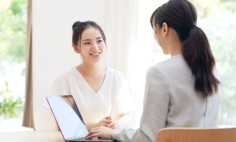 Two women have a friendly conversation, one using a laptop. Bright, airy setting.