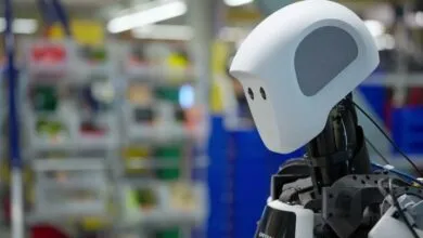 Close-up of a white robot head with sensors, in a blurred factory setting.
