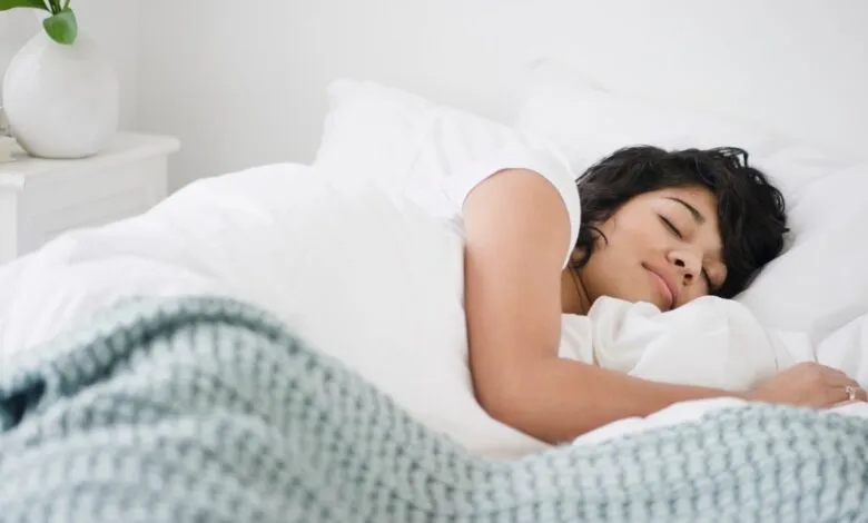 Woman peacefully sleeping in bed, covered by white sheets and a light teal blanket.