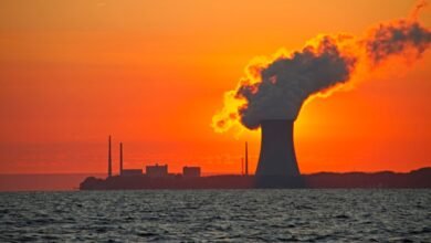 Silhouetted power plant at sunset, smoke billowing against a fiery orange sky over dark water.