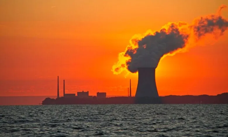 Silhouetted power plant at sunset, smoke billowing against a fiery orange sky over dark water.