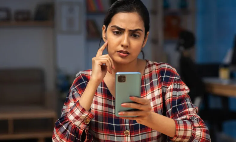 Woman looks thoughtfully at her smartphone, hand to chin, in a home office.