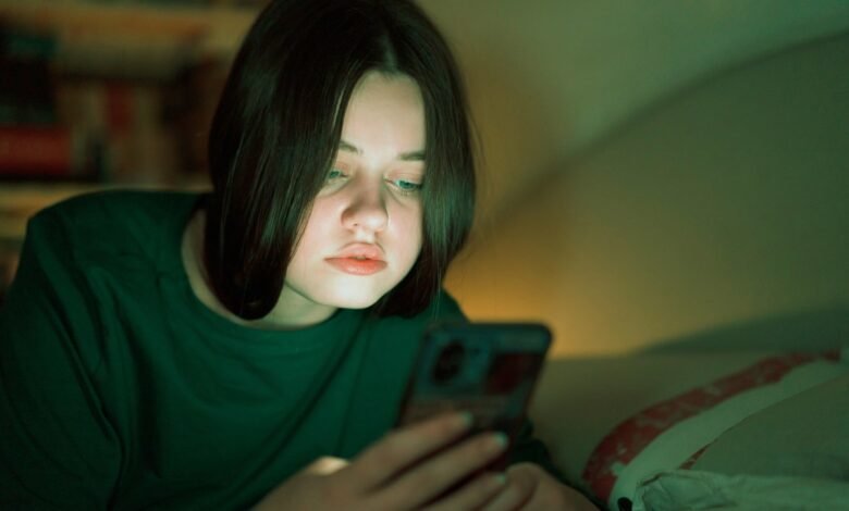 Teenage girl in green shirt lies in bed at night, intently looking at her smartphone screen illuminated by its light.