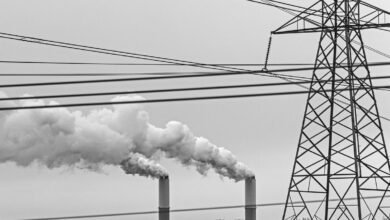 Black and white image of industrial smokestacks emitting smoke, with a power line tower and wires.