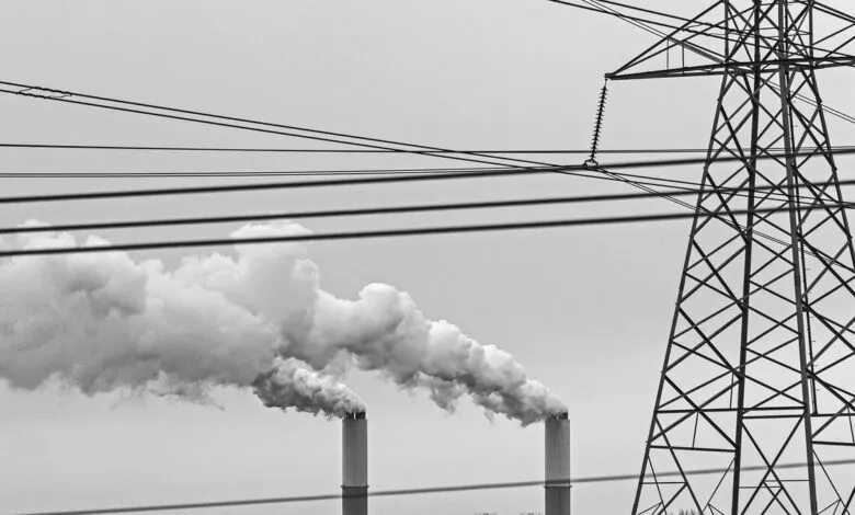Black and white image of industrial smokestacks emitting smoke, with a power line tower and wires.