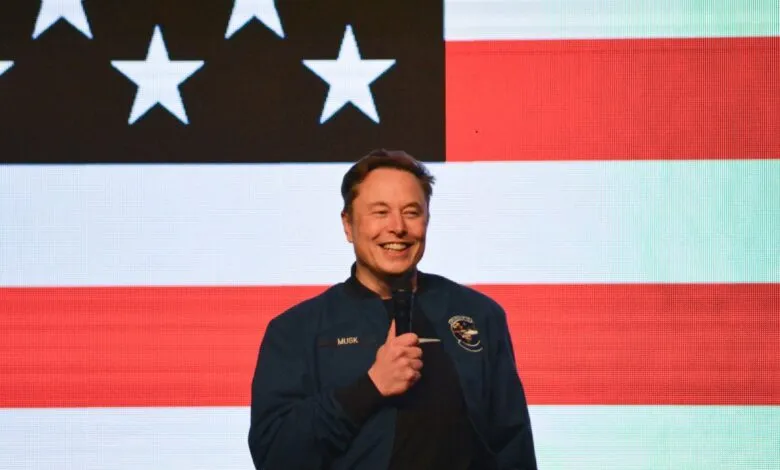 Smiling Elon Musk gives a thumbs-up, speaking in front of an American flag backdrop.