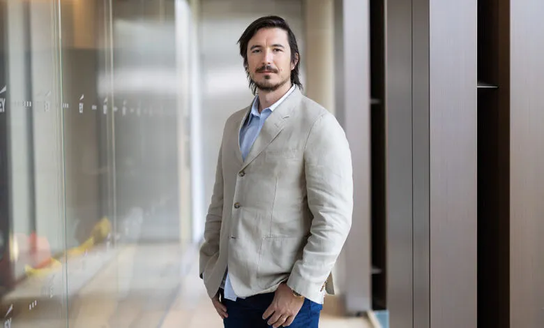 Vlad Tenev, Robinhood CEO, in a beige blazer, stands in a modern office hallway.