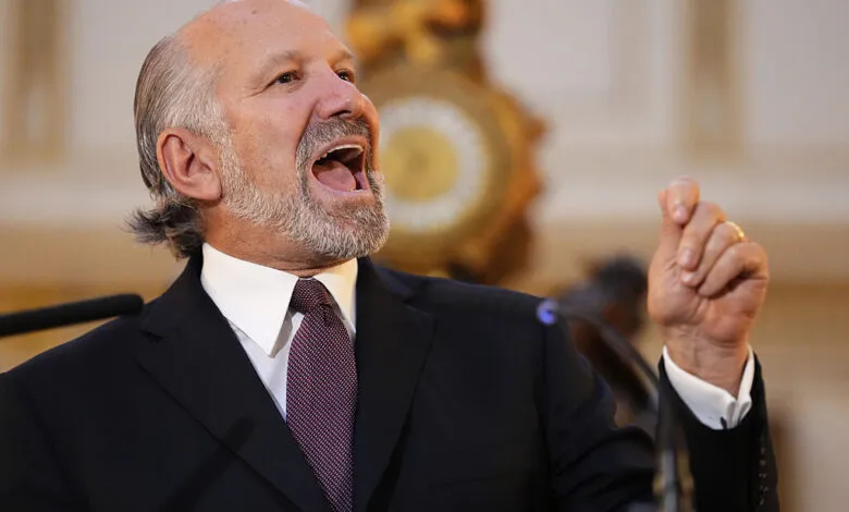 Man in a suit speaking passionately with his mouth open and hand raised.
