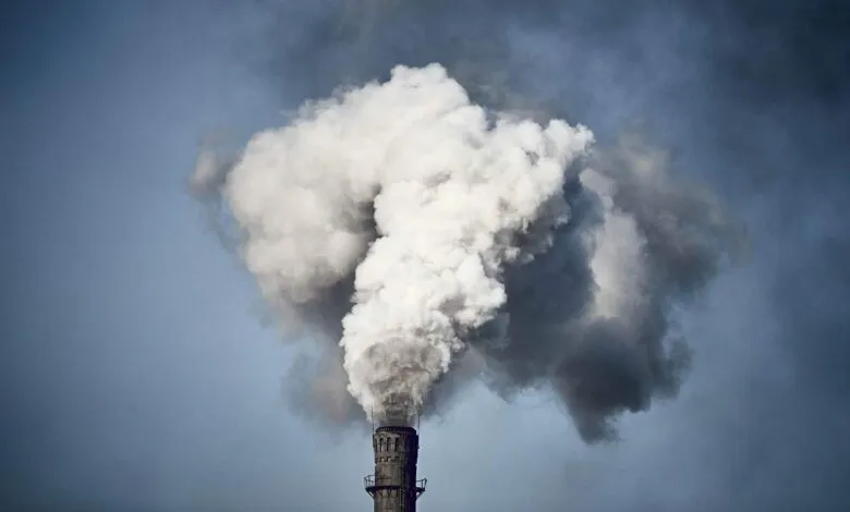 Industrial smokestack emitting a large plume of gray and white smoke against a muted blue sky.
