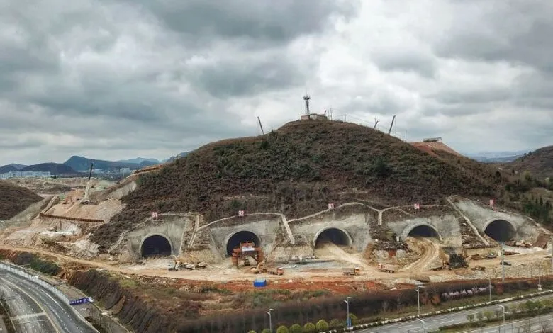 Multiple tunnel entrances carved into a hillside under construction, with heavy machinery visible.