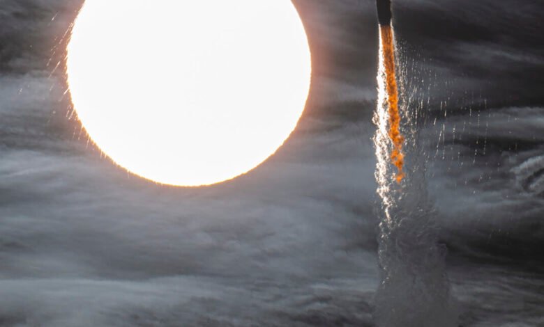 Rocket launch plume against a bright sun and cloudy sky.