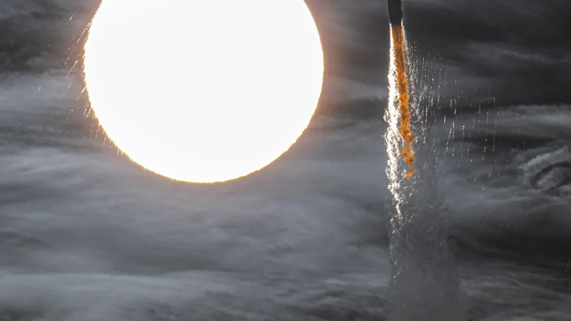 Rocket launch plume against a bright sun and cloudy sky.