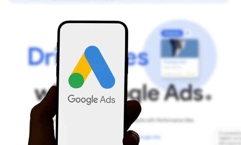 Close-up of a smartphone displaying the Google Ads logo. The background shows a blurred Google Ads webpage.