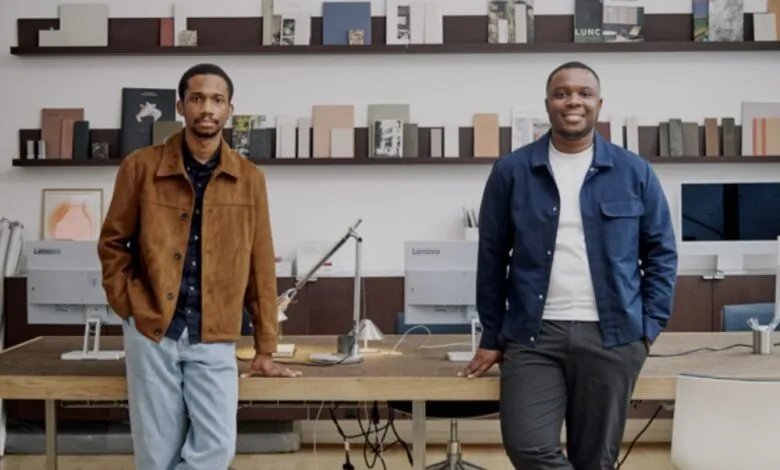 Two men stand in a modern office, leaning on a desk. Neutral color palette, bookshelves in background.