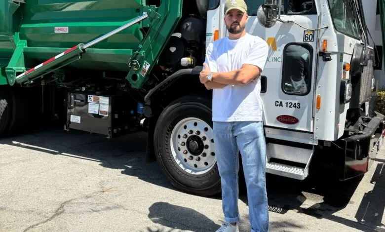 Man in white t-shirt and jeans stands in front of a green and white garbage truck.