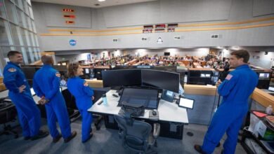 Astronauts in blue suits briefing a large audience in a NASA mission control room.