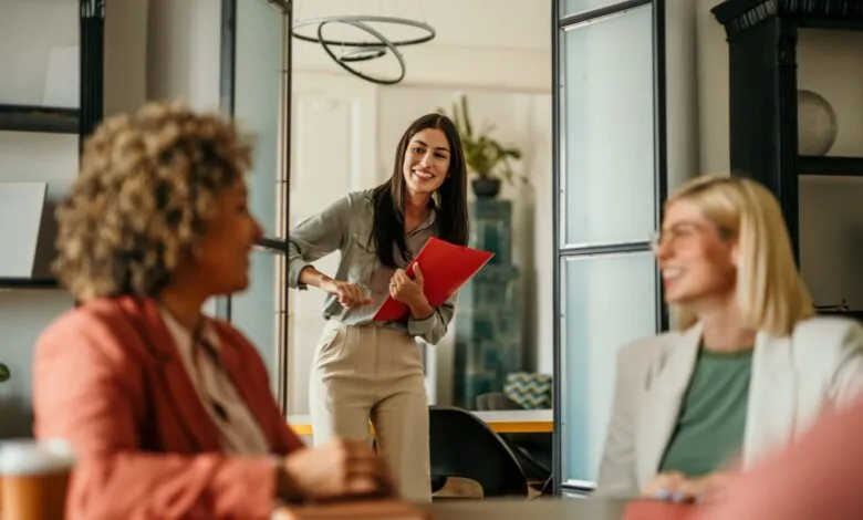 Smiling businesswoman enters a meeting, holding a red folder; colleagues await.