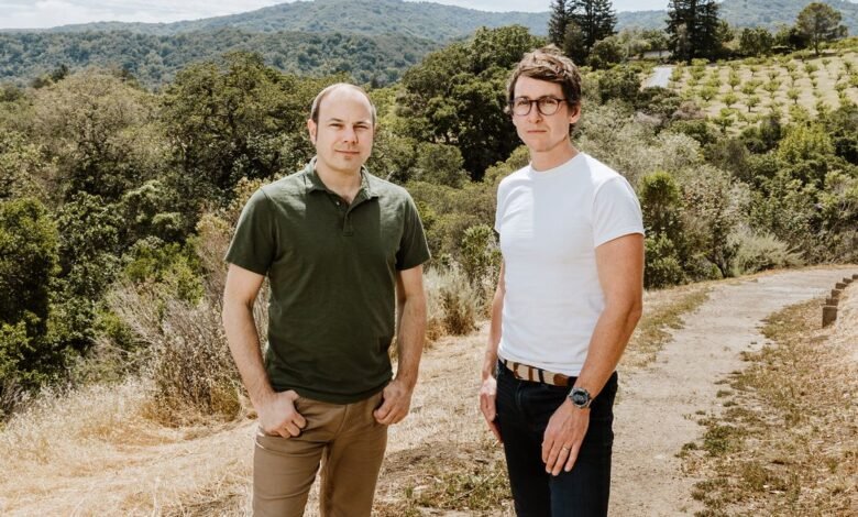 Two men stand outdoors, one in a green shirt and the other in white, against a backdrop of rolling hills and trees.