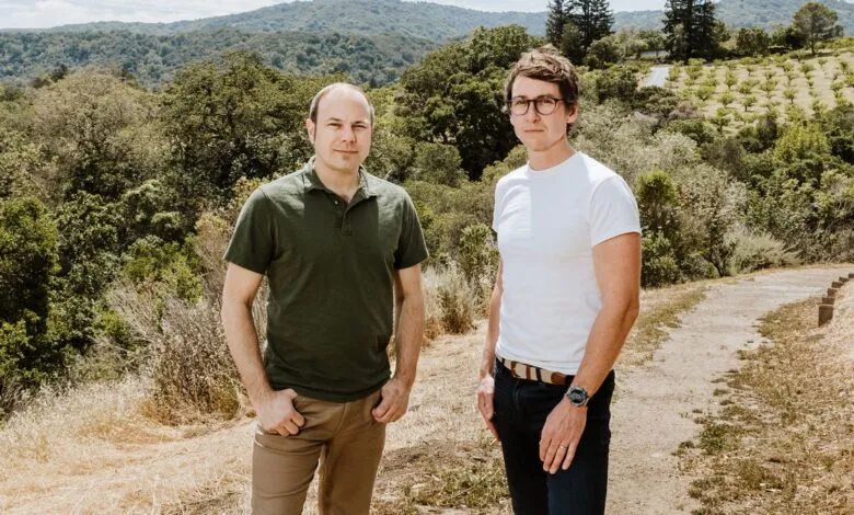 Two men stand outdoors, one in a green shirt and the other in white, against a backdrop of rolling hills and trees.