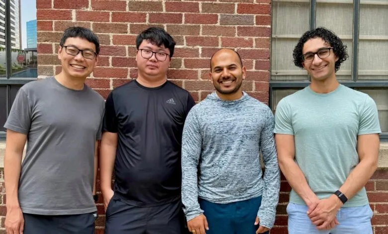 Four smiling men stand against a brick wall. They wear casual clothing.