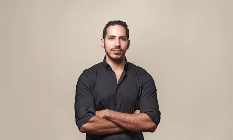 Portrait of a man with arms crossed, wearing a black shirt against a beige background.