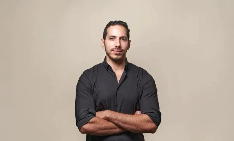 Portrait of a man with arms crossed, wearing a black shirt against a beige background.