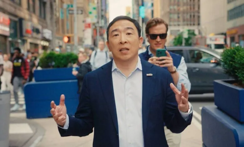 Andrew Yang, in a navy suit, speaks passionately on a busy NYC street, while a person films him.