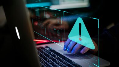 Person's hand on laptop touchpad with a teal warning alert overlay. Cybersecurity risk concept.