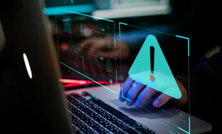 Person's hand on laptop touchpad with a teal warning alert overlay. Cybersecurity risk concept.