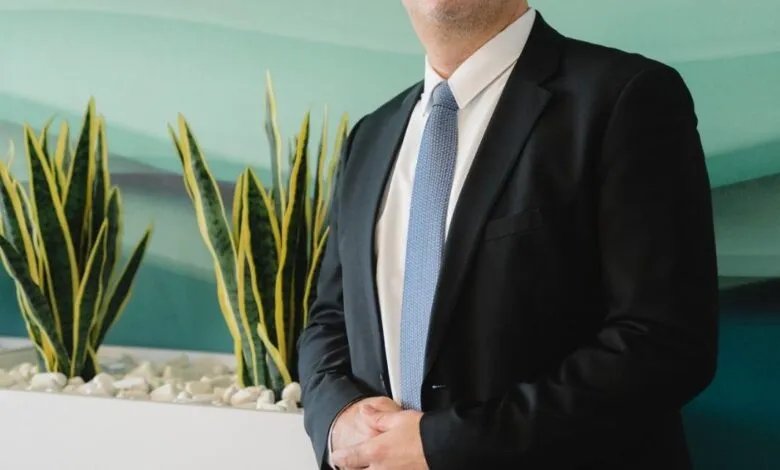 Portrait of Raif Abou Diab, a man in a suit, smiling subtly against a teal wall.