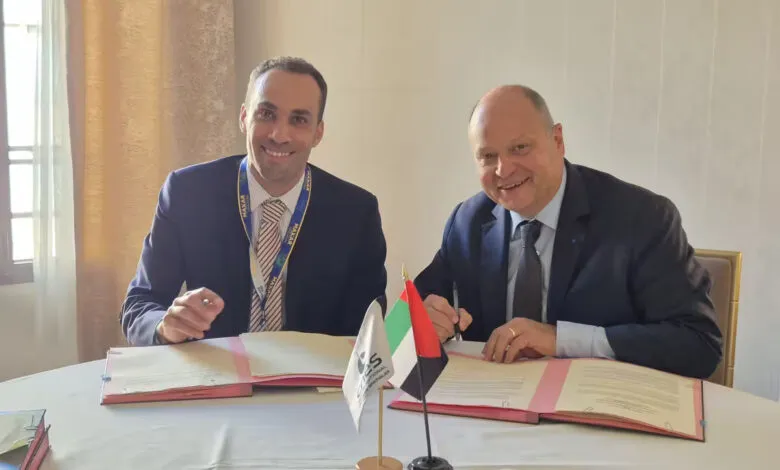 Two men in suits smiling, signing a document at a table with flags. Business agreement.