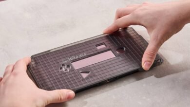 Hands carefully placing a translucent gray gridded device on a light gray surface. The device has a small pink rectangle and several cutouts.