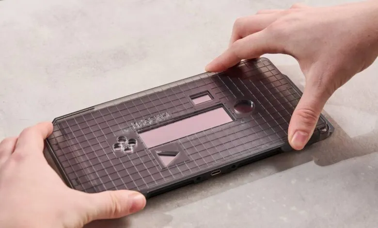 Hands carefully placing a translucent gray gridded device on a light gray surface. The device has a small pink rectangle and several cutouts.