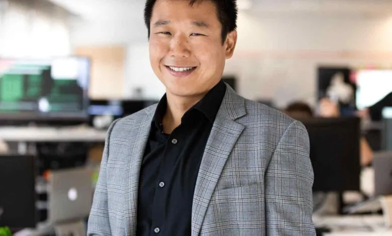 A smiling Asian man in a grey plaid suit jacket and black shirt stands in an office.