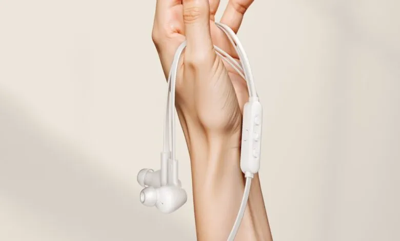 A hand holds white wired earbuds with a control module. The earbuds appear to have noise-canceling capabilities.