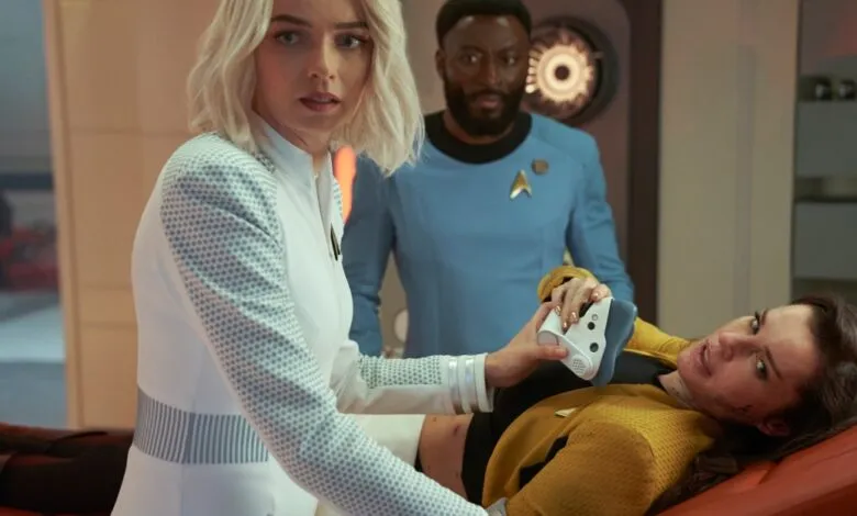 Woman in white uniform uses medical device on injured woman in yellow uniform, man in blue uniform watches.