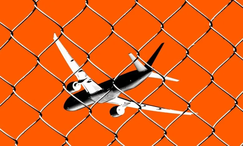 Monochrome airplane seen through a chain-link fence against a bright orange background.