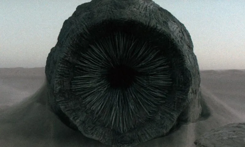 Mysterious dark object resembling a giant eye, partially buried in sand under a hazy sky.